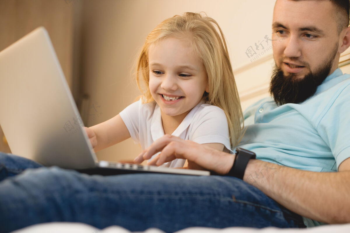 男人小胡子爸爸和他的小女儿靠在家里的床上看着笔记本电脑屏幕肖像教育爸爸