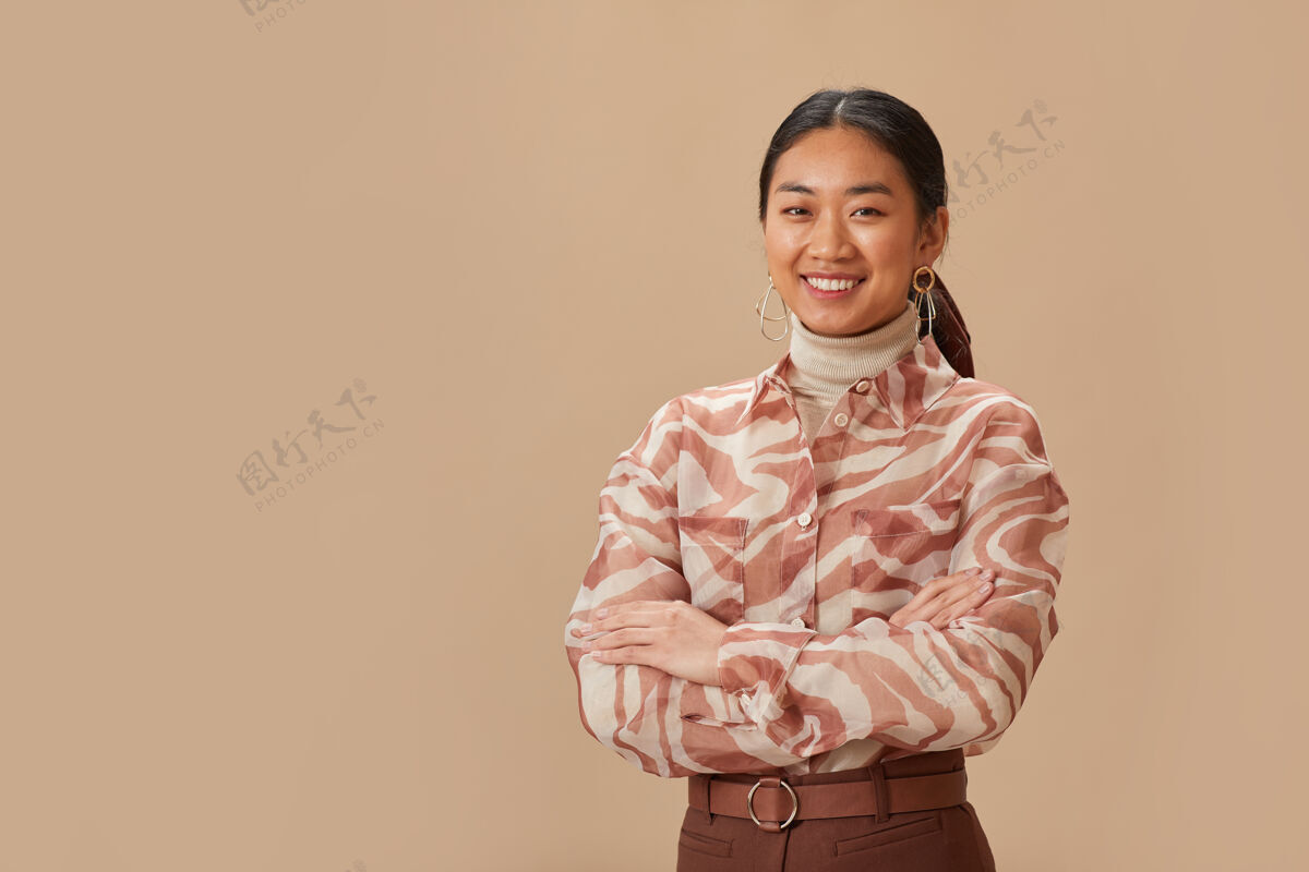 自信身着优雅服装的亚洲美女的肖像画 双臂交叉 面带微笑地站在米色的墙上双臂交叉职业休闲服