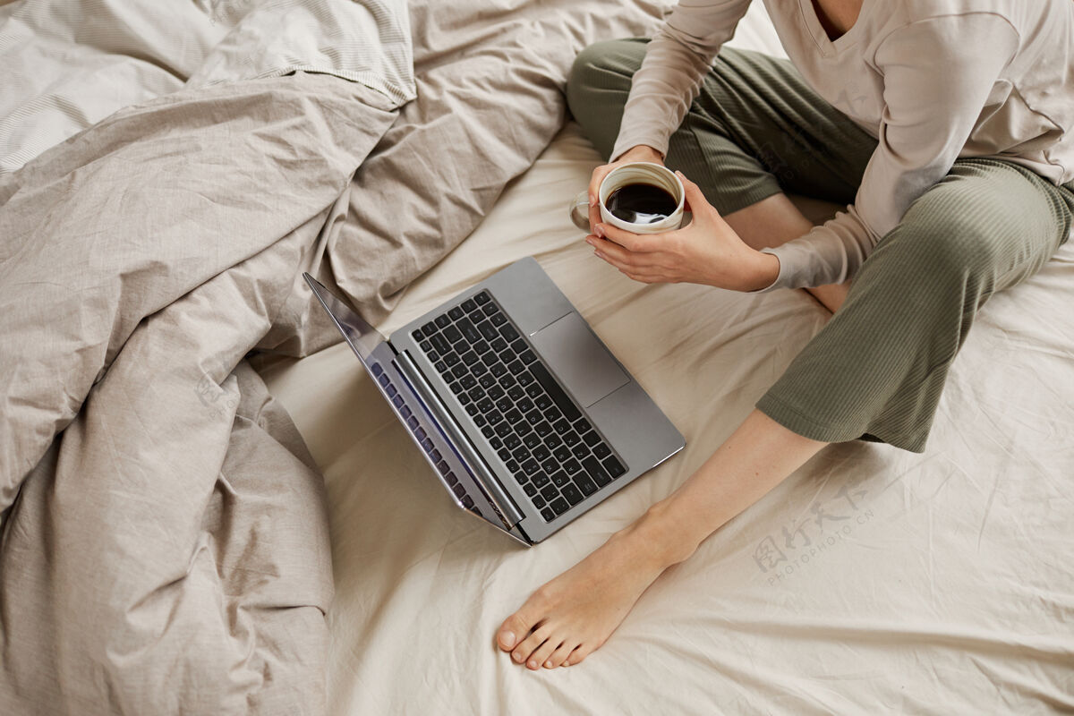 家庭生活坐在床上喝咖啡 在家里用笔记本电脑看东西的年轻女子的特写镜头观看杯子通讯