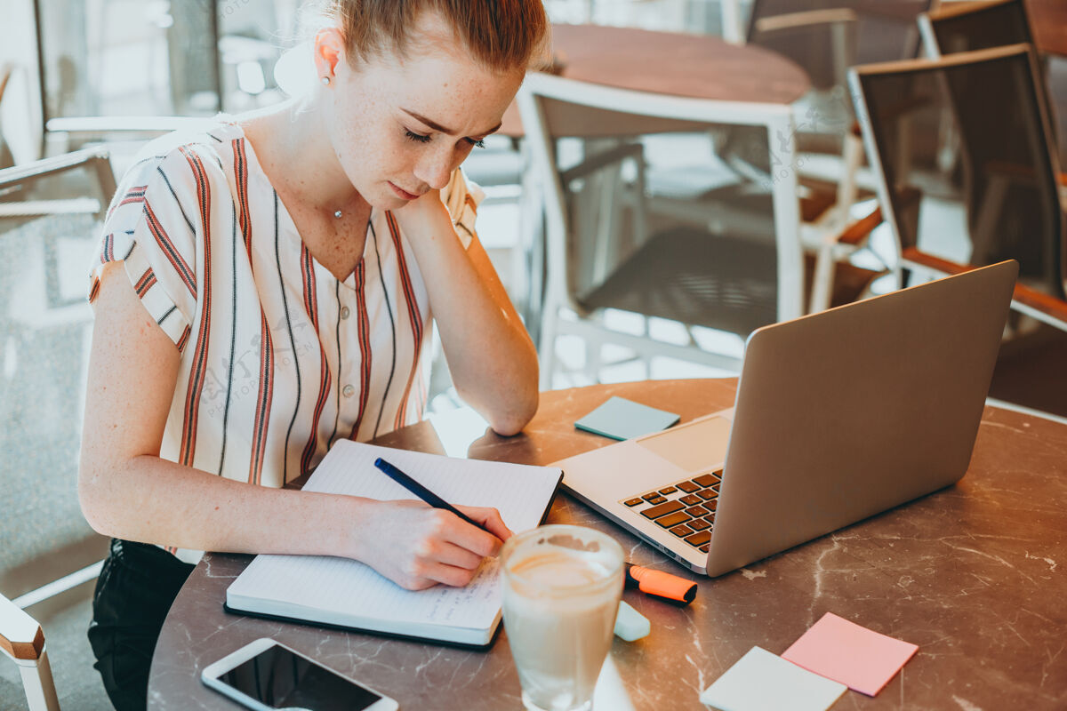 社交媒体上图是一位年轻的商务女性 红发 长着雀斑 在咖啡馆的户外笔记本上做笔记 桌上放着一台笔记本电脑笔记在线网络