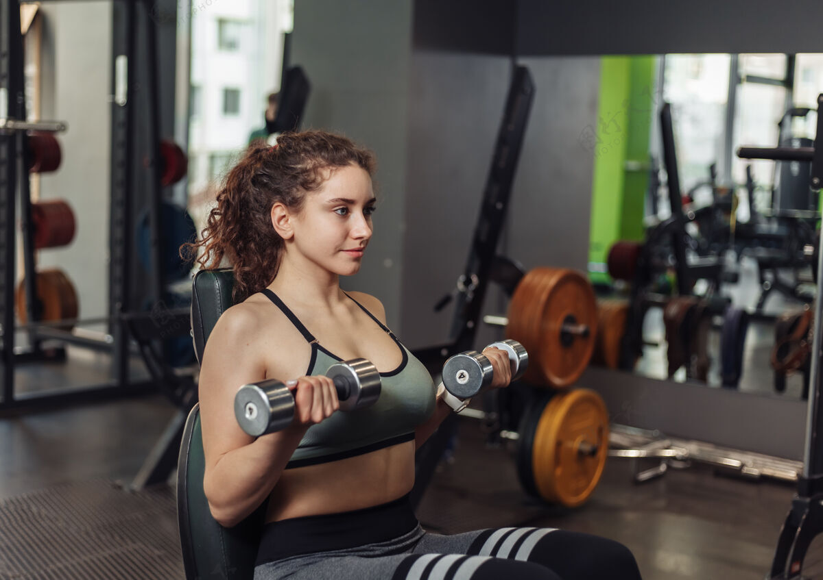 活跃年轻健康的女子坐在健身房的长凳上练习举起哑铃健身坐着形状