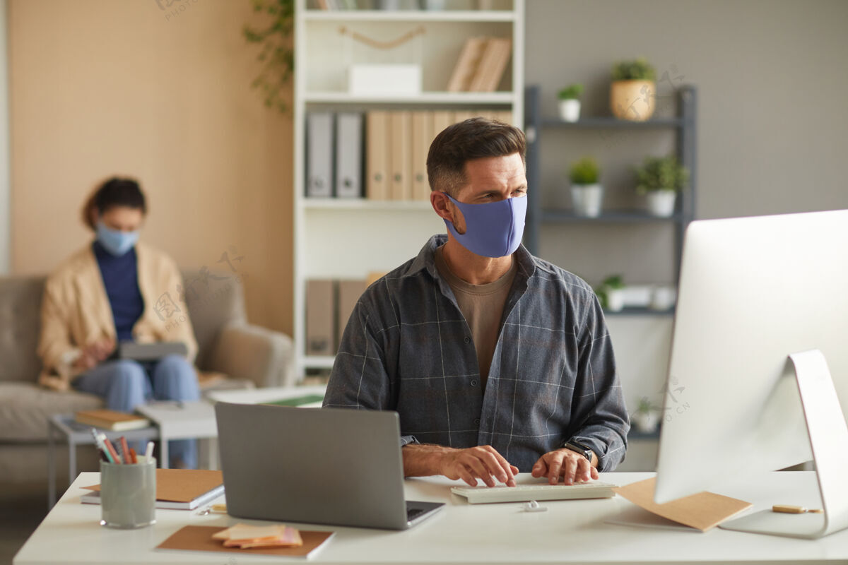 办公桌戴着防护面具的商人和同事坐在办公室的墙上 在桌子旁的电脑上工作职业笔记本电脑