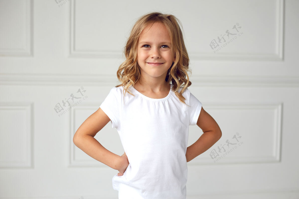 时尚正面肖像是一个小女孩微笑着看着镜头 穿着白色t恤 手放在腰上快乐可爱金发