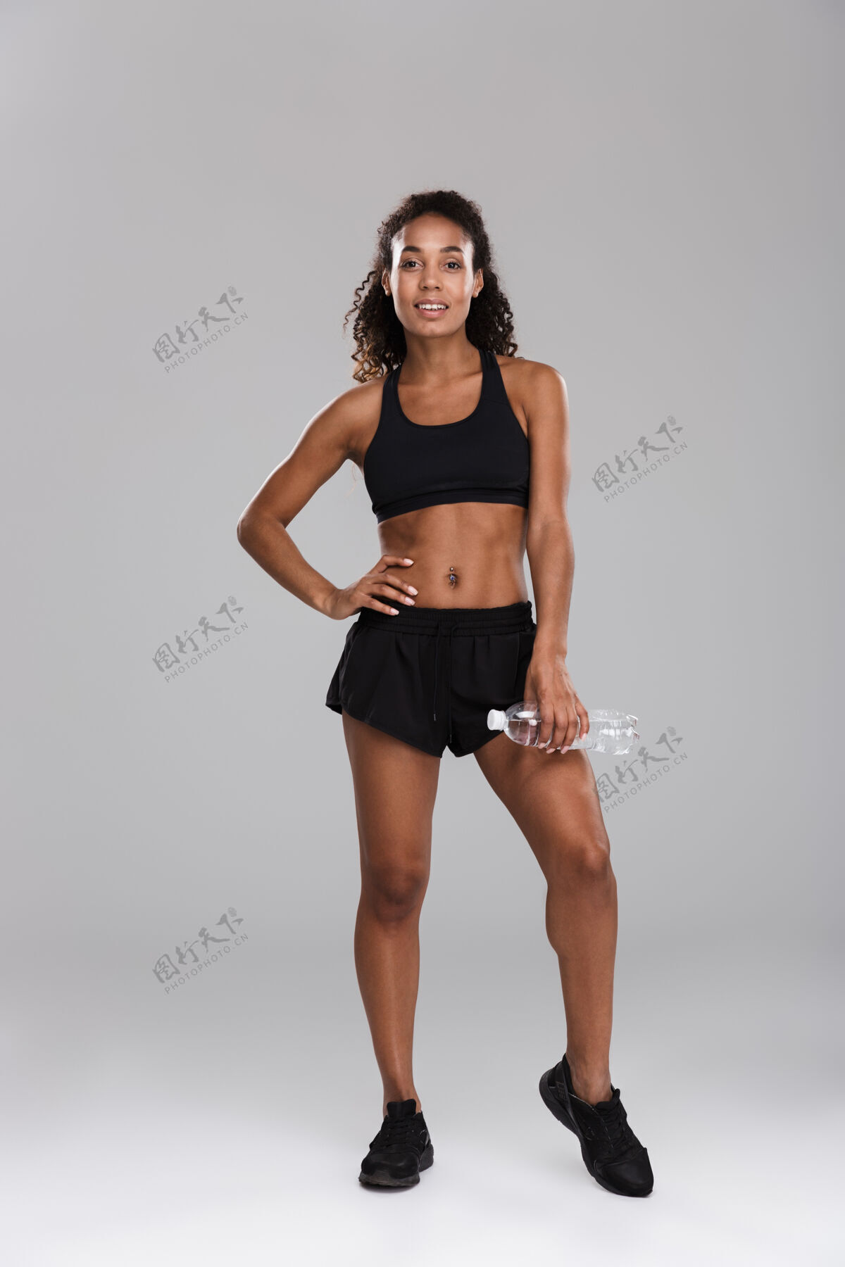 美国一位微笑的年轻非洲女运动员的全长肖像 她在灰色的墙壁上独自训练后休息 手里拿着装满水的瓶子饮食微笑长度