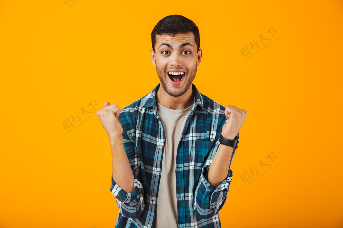 成功一个穿着格子衬衫的快乐的年轻人孤零零地站在橘色的墙上庆祝兴奋年轻健身