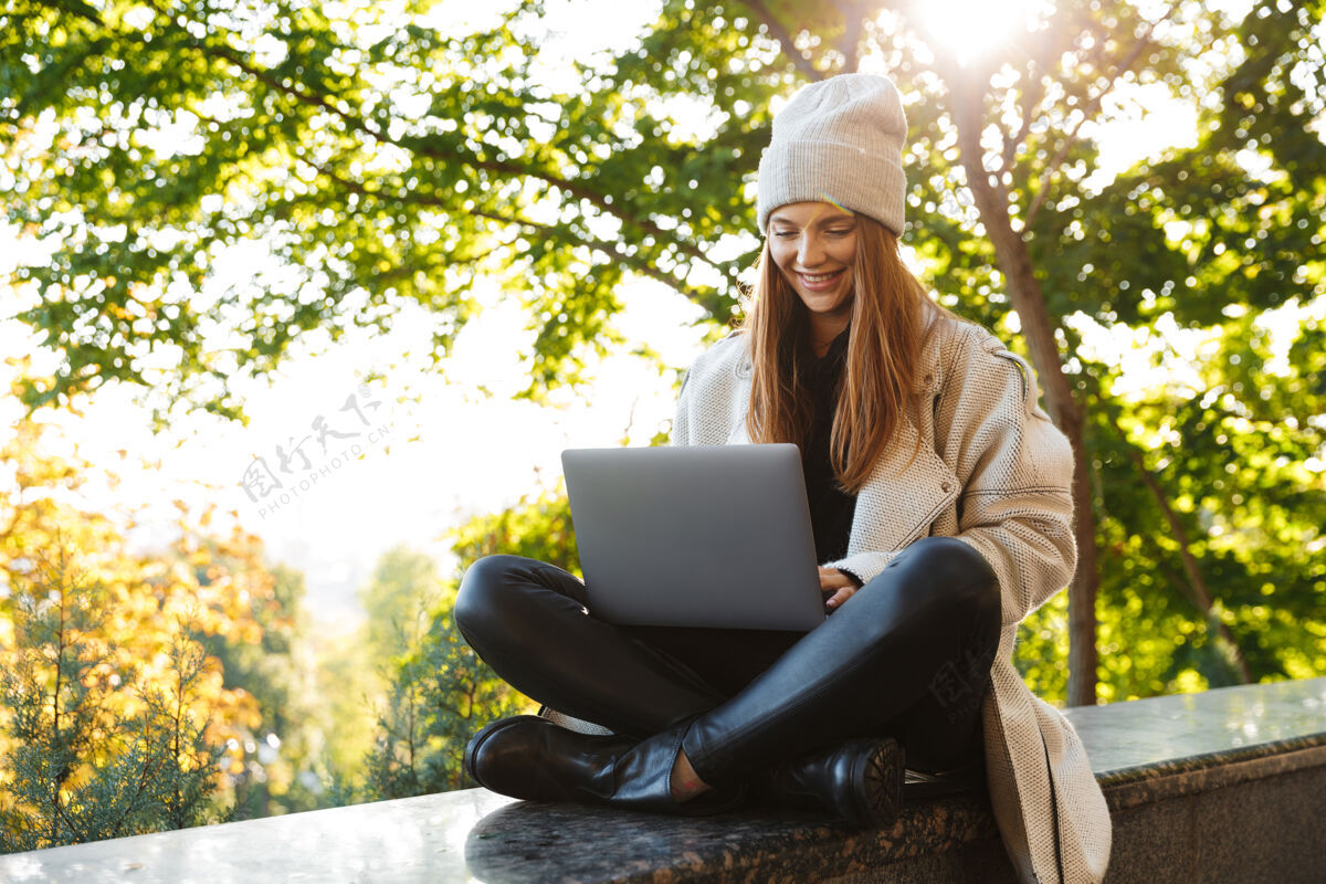 笔记本电脑美丽的年轻女子穿着秋大衣 戴着秋帽坐在户外 用笔记本电脑电脑类型成功