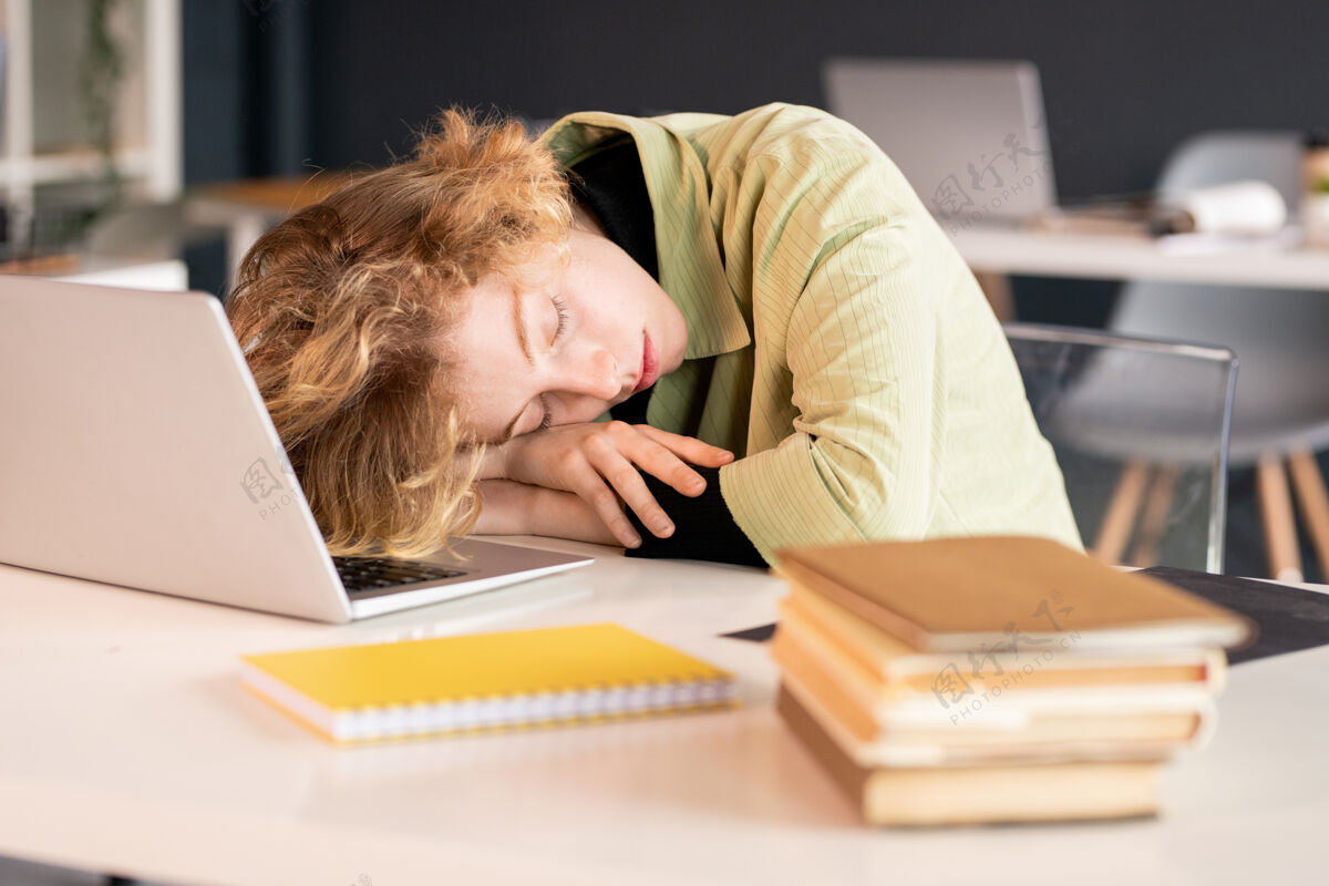 过度工作年轻精疲力尽的女学生或办公室经理 在笔记本电脑前小睡时 头靠在手上 旁边有一堆书室内男士情绪紧张