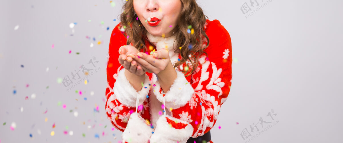 明亮圣诞节 人们的概念-穿圣诞套装的女人在墙上吹五颜六色的亮片西装女人年轻