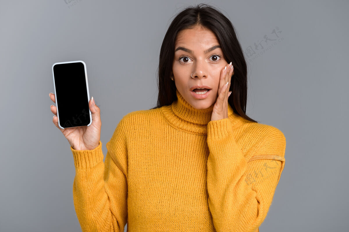 人一个震惊的随意的年轻女子的肖像隔离在灰色的墙上 显示手机的空白屏幕震惊成人显示