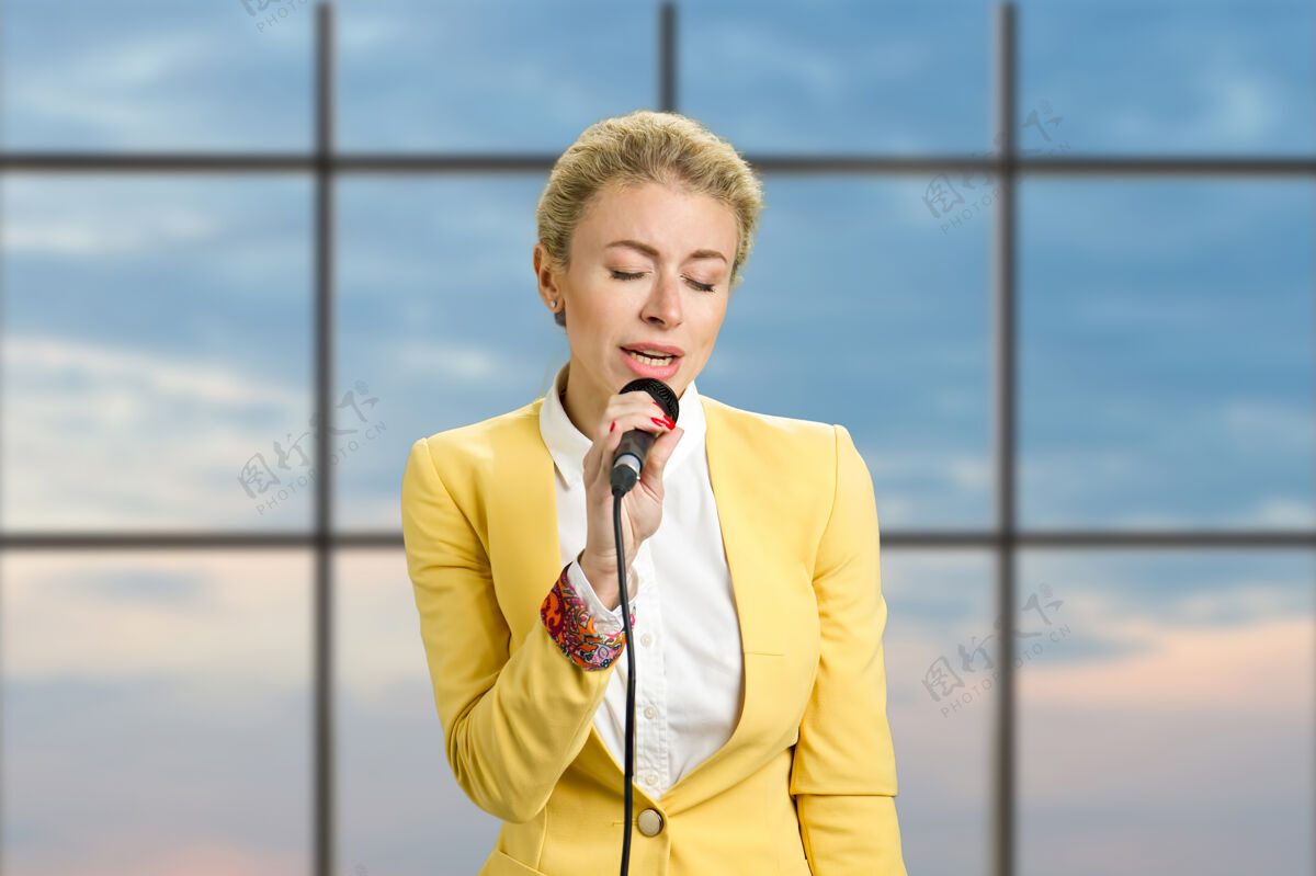 演示戴着麦克风闭着眼睛的年轻女士漂亮的金发女商人对着办公室窗户上的麦克风说话商务女性封闭歌曲