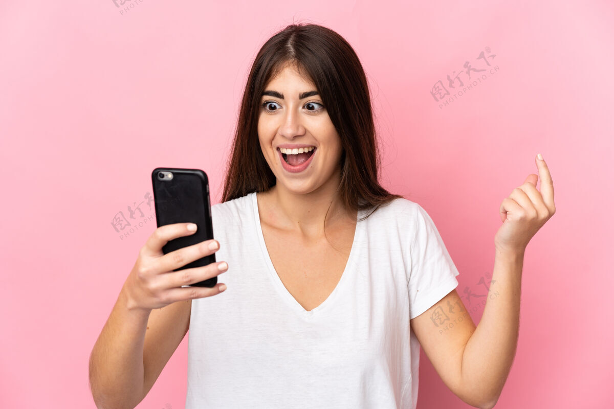 担心年轻的白人妇女孤立在粉红色背景下 一边用手机一边看着相机 表情惊讶黑发女性沟通