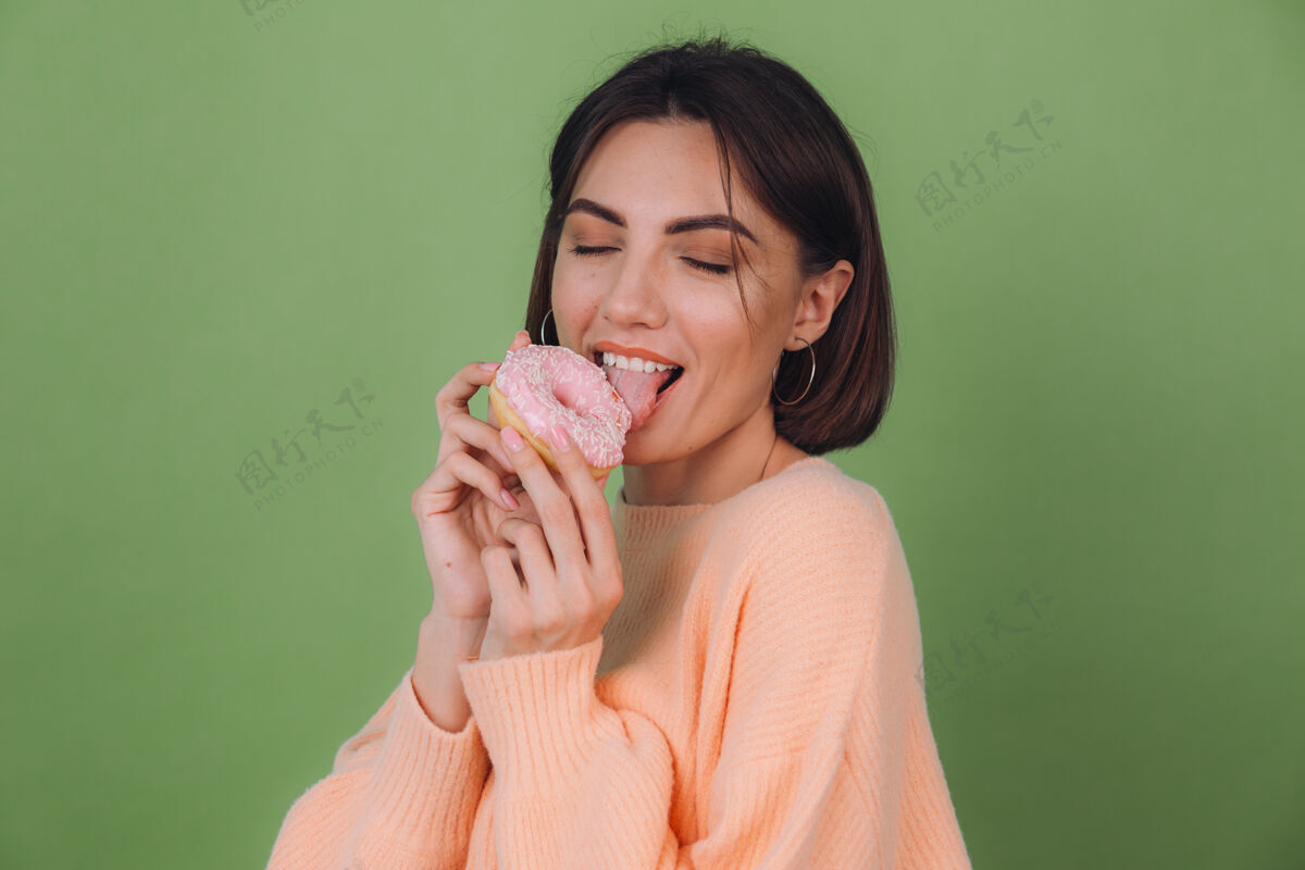 食物年轻时尚的女人穿着休闲的桃色毛衣孤立地站在绿橄榄色的墙上舔着粉色的甜甜圈复制空间黑发饥饿甜点
