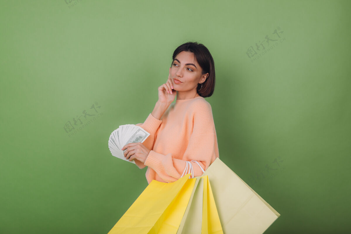 聪明穿着休闲桃色毛衣的年轻女子孤零零地站在绿橄榄墙上 手里拿着100美元钞票和购物袋的扇子 想着积极微笑的复制空间时尚立场壁板
