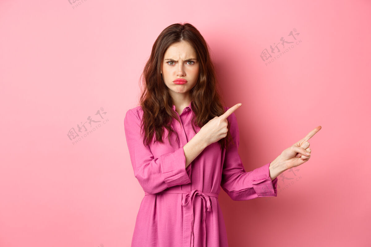快乐美丽和时尚穿着时髦衣服的愤怒的女士皱眉不高兴 评判某人 用手指指着不好的宣传文案 站在粉红色的墙上候选人兴奋时尚