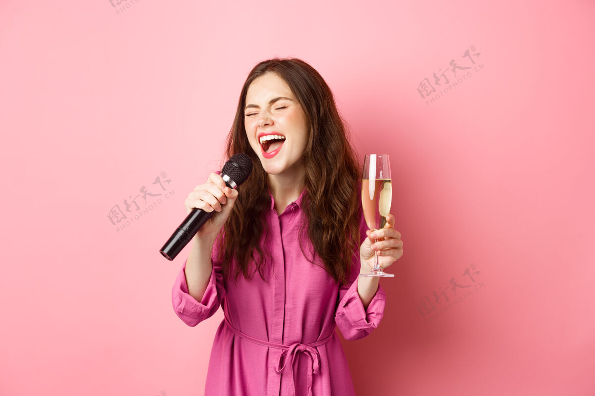 香槟庆祝和节日的概念派对女士玩得开心 喝香槟 唱卡拉OK 拿着麦克风 快乐地站在粉红色的墙上麦克风饮酒水平