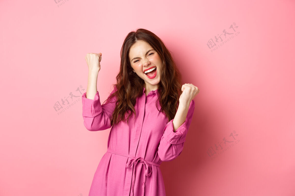 情绪美丽的微笑的女人带着快乐和兴奋的脸尖叫 说是的 做拳头泵 胜利和感觉像冠军 胜利 站在粉红色的墙上咧嘴笑请乐观