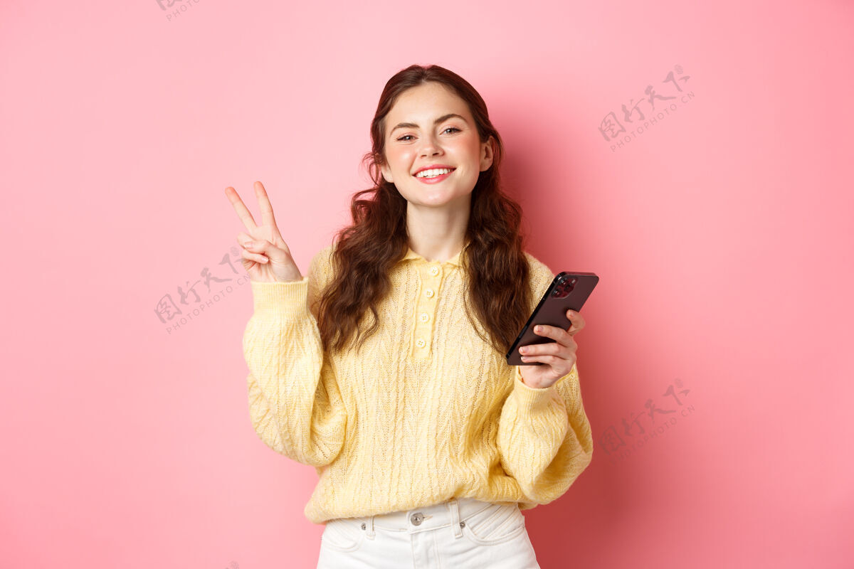 成人科技和网上购物快乐的年轻女子微笑着高兴 手持智能手机 显示v型标志 看着相机 站在粉红色的墙上年轻在线微笑