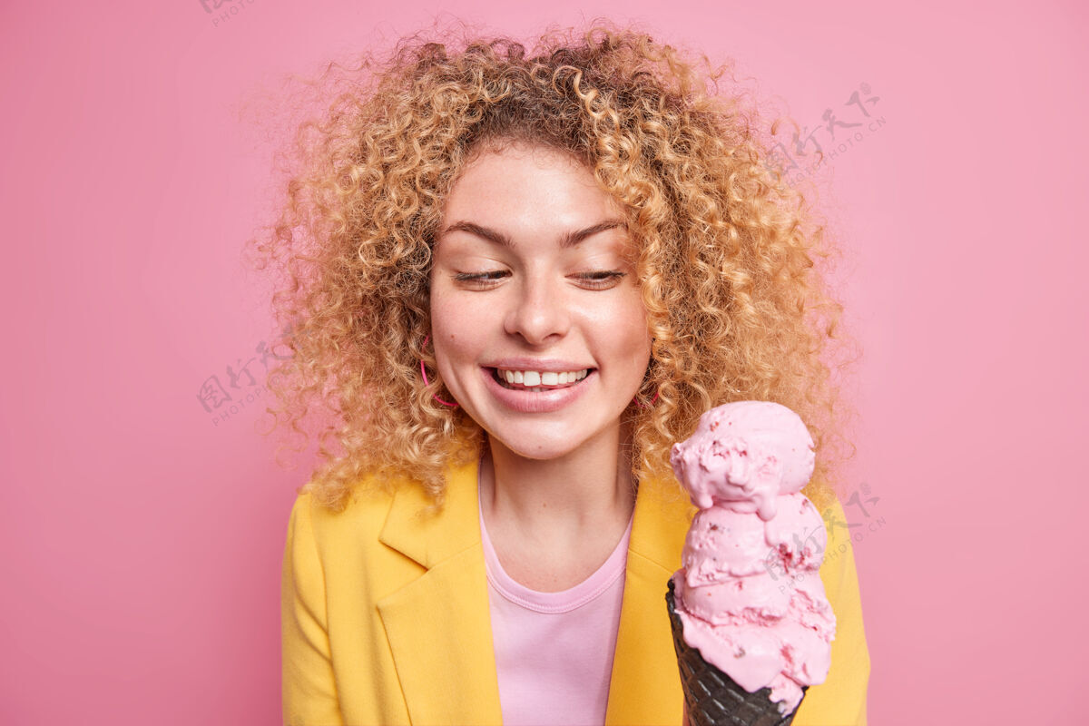 风味室内拍摄的漂亮开朗的欧洲女人开心地微笑着看着开胃的粉红色冰淇淋在黑色华夫饼穿着黄色夹克自然卷发隔离在玫瑰色的墙壁惊人欢乐欧式