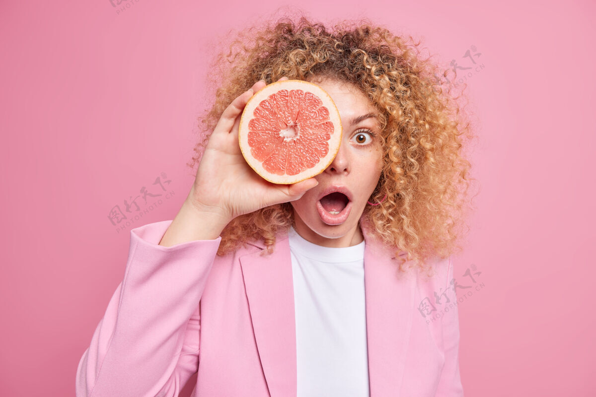 卷发惊讶的卷发欧洲女人用葡萄柚片遮住眼睛 穿着时尚的衣服张开嘴 隔着粉色的墙壁夏日茶点柑橘类水果概念罗西水果果汁