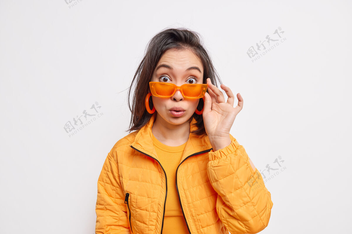 女孩令人惊讶的时尚黑发亚洲年轻女子戴着时髦的橙色太阳镜夹克和耳环 在白色墙壁上对一些令人惊讶的孤立的东西做出反应风格和时尚概念成人惊喜夹克