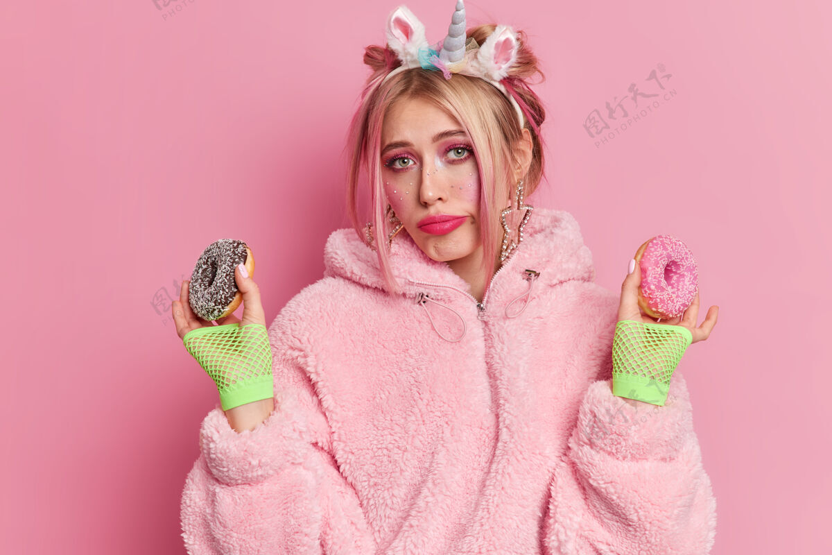 金发苦恼不快乐的女人戴着独角兽头带保暖外套和运动手套拿着两个釉面甜甜圈尽量避免吃有害食物保持节食感觉诱惑不满年轻美味