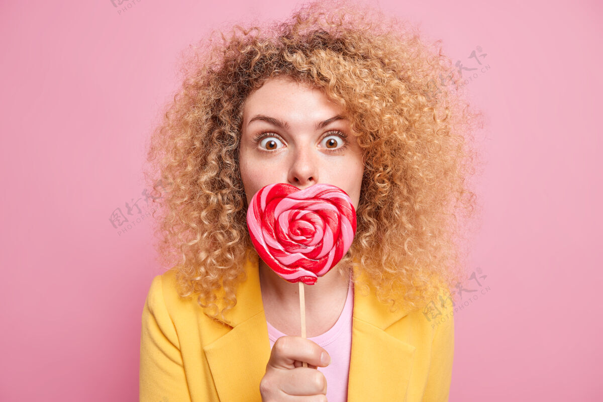 心惊讶的卷发年轻女子用心形糖果捂住嘴吃甜食拿着棒棒糖穿着黄色夹克隔离在粉红色的墙上糖太多不好甜食女性肖像青少年