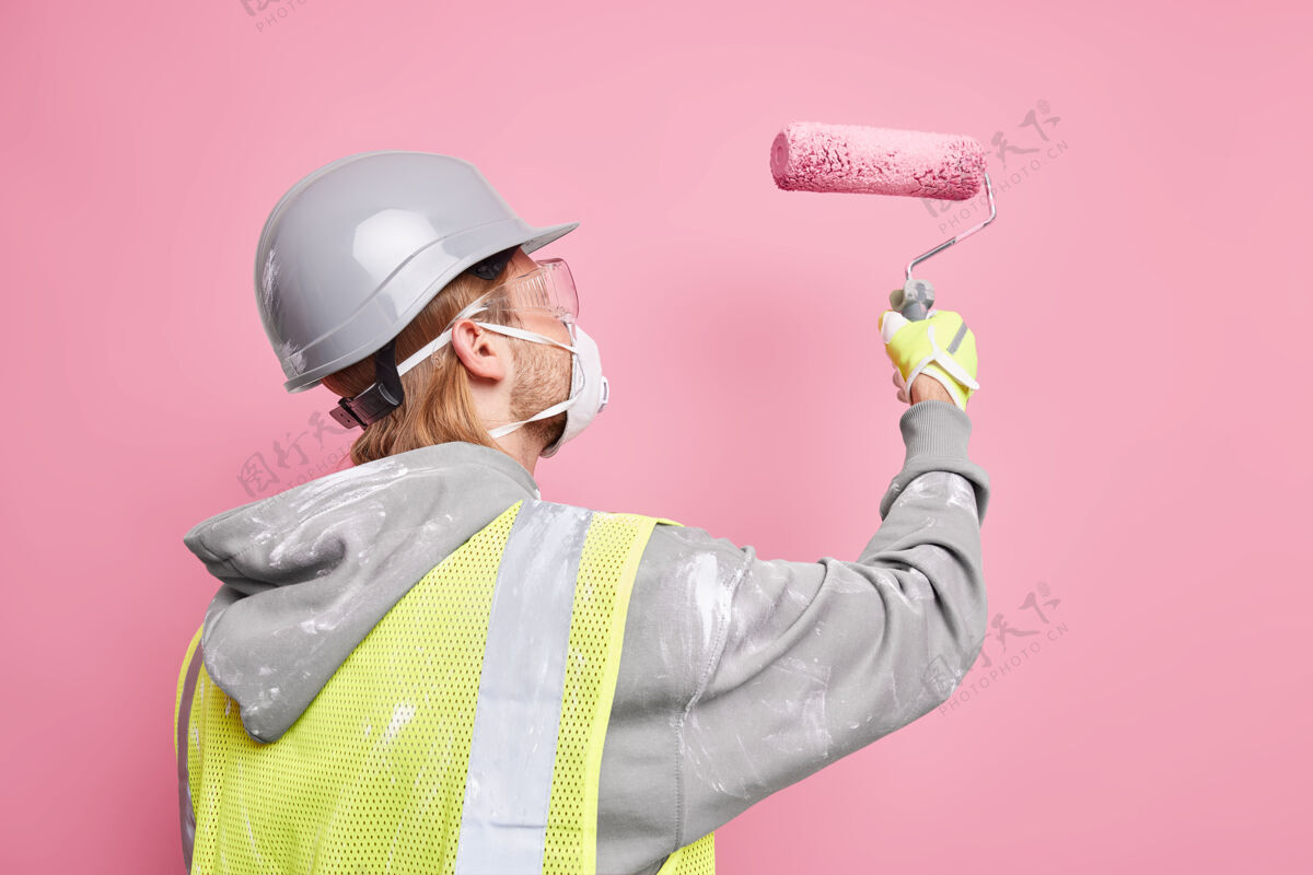安全帽穿著安全制服的忙碌杂工手持油漆辊戴著防护面罩在粉红色墙壁上努力隔离工作维修和翻新概念维修工人支架眼镜室内