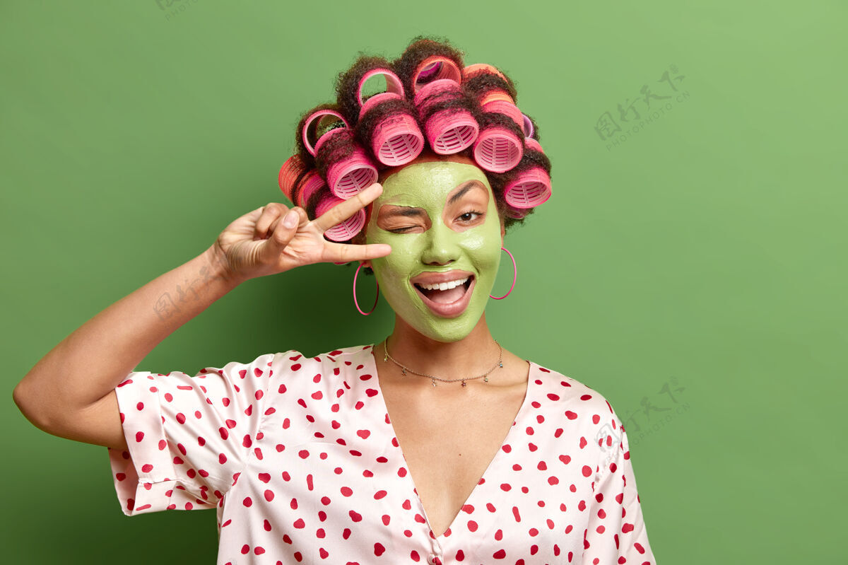 清洁积极向上的主妇喜欢花时间关心自己塑造v字型眨眼有欢快的表情应用有效的绿色面膜做发型穿圆点睡衣面部模特胜利