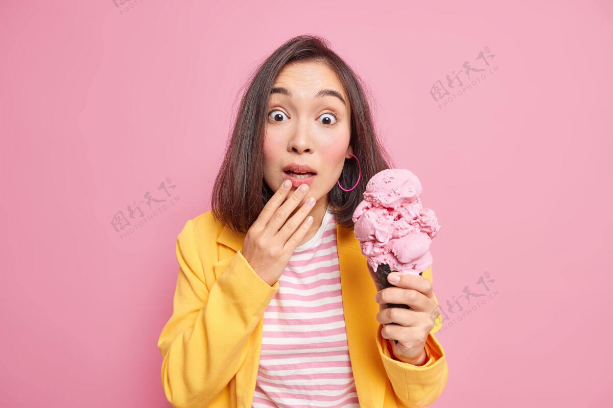 人类一位惊讶的亚洲年轻女子 深色头发 穿着条纹t恤和黄色夹克 拿着美味的冰淇淋隔着粉色的墙人和情感的概念自然美味美味