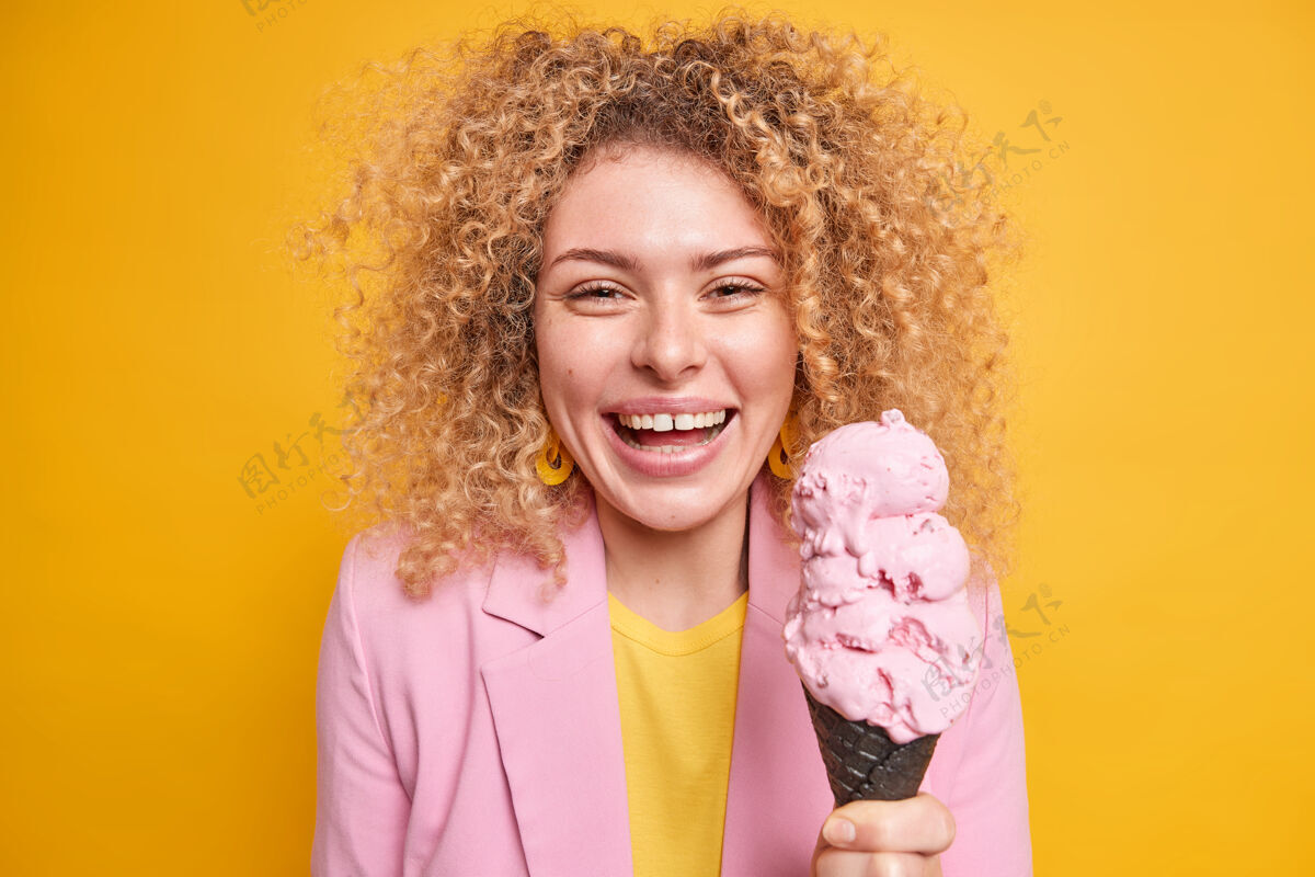 糖一位留着卷发的漂亮女人的肖像她拿着锥形冰激凌 在夏天和朋友们在公园里散步 被隔离在黄色的墙壁上女模特吃着美味的甜点室内夹克味道