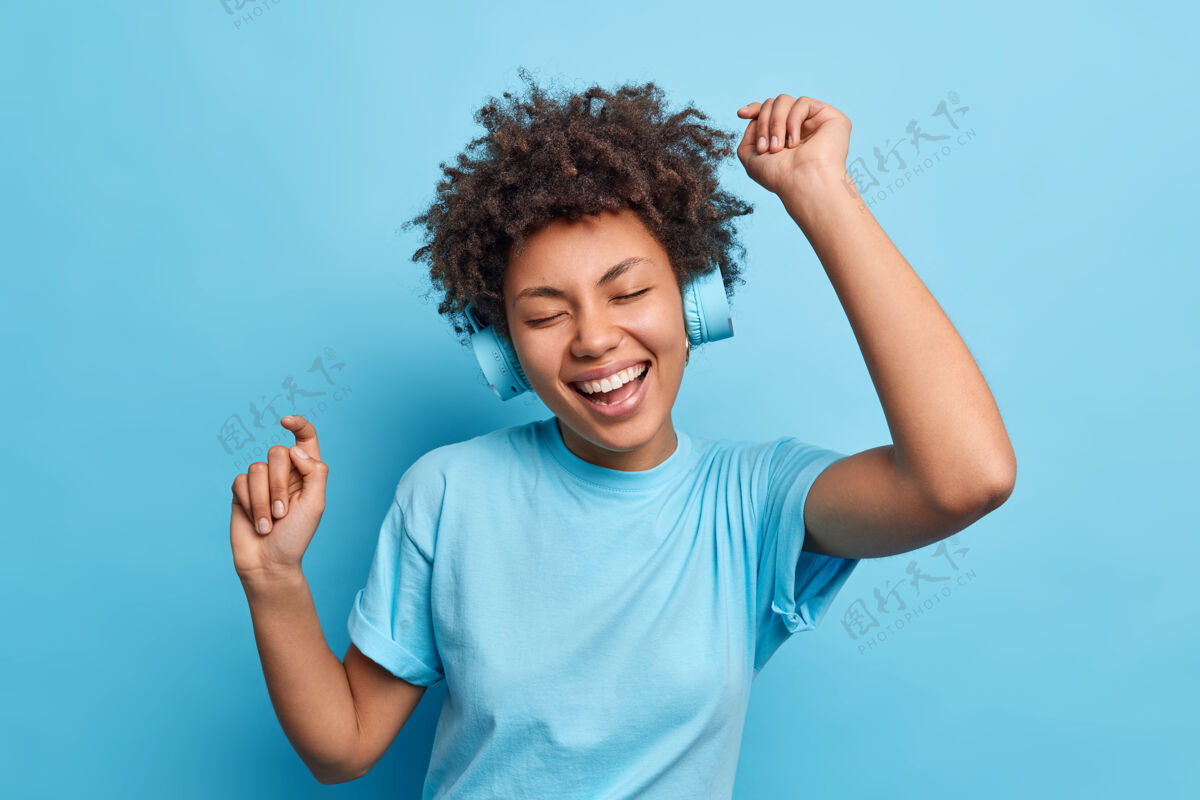 单色快乐放松的非洲裔美国女孩喜欢最喜欢的播放列表通过可怜的耳机听音乐举起手臂穿着休闲隔离在蓝色的墙壁人们的爱好和生活方式的概念年轻卷发站