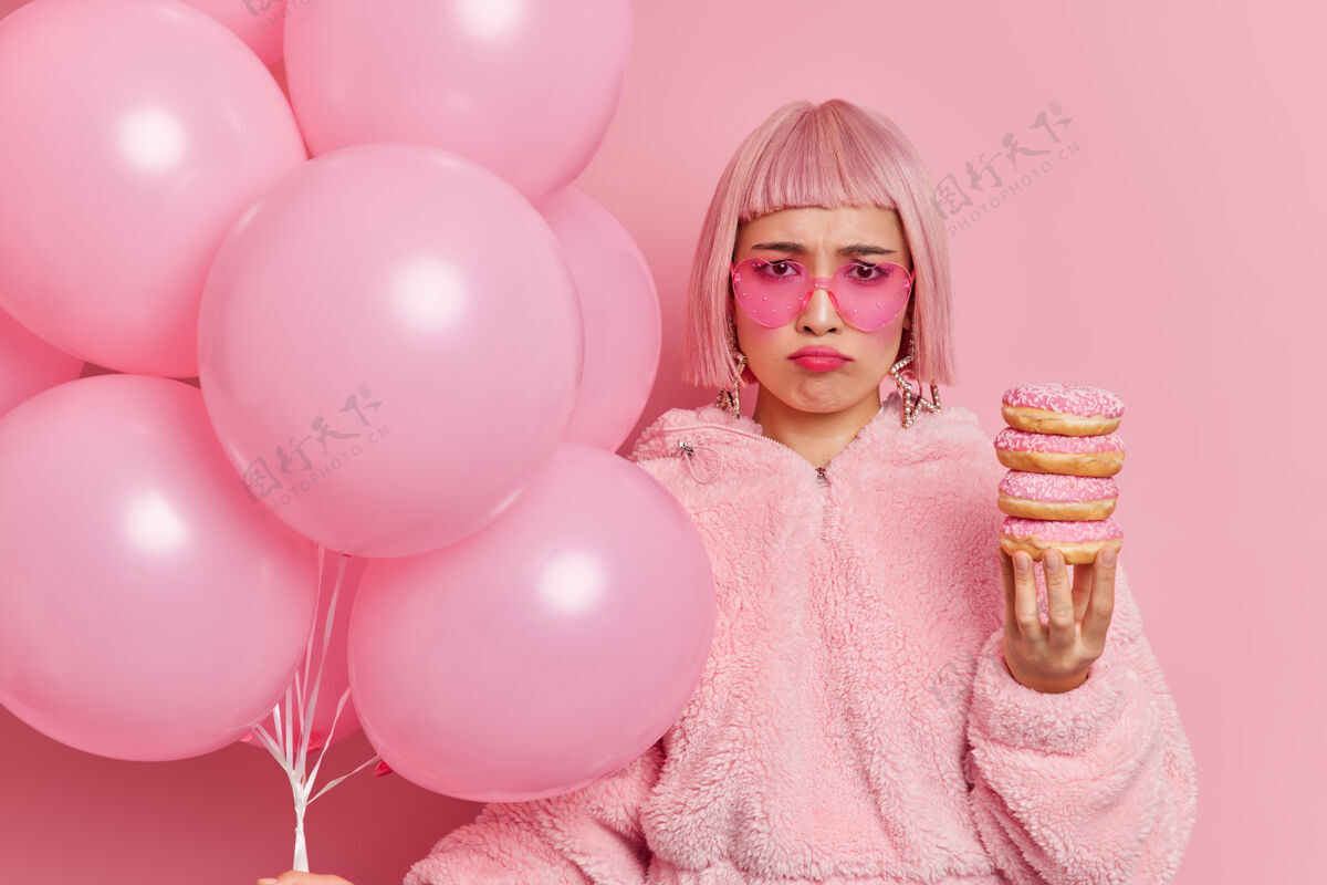 粉色不满节俭的亚洲女人戴着时髦的太阳镜心情不好 独自庆祝生日时手里拿着一堆甜甜圈和充气气球人皮毛时尚