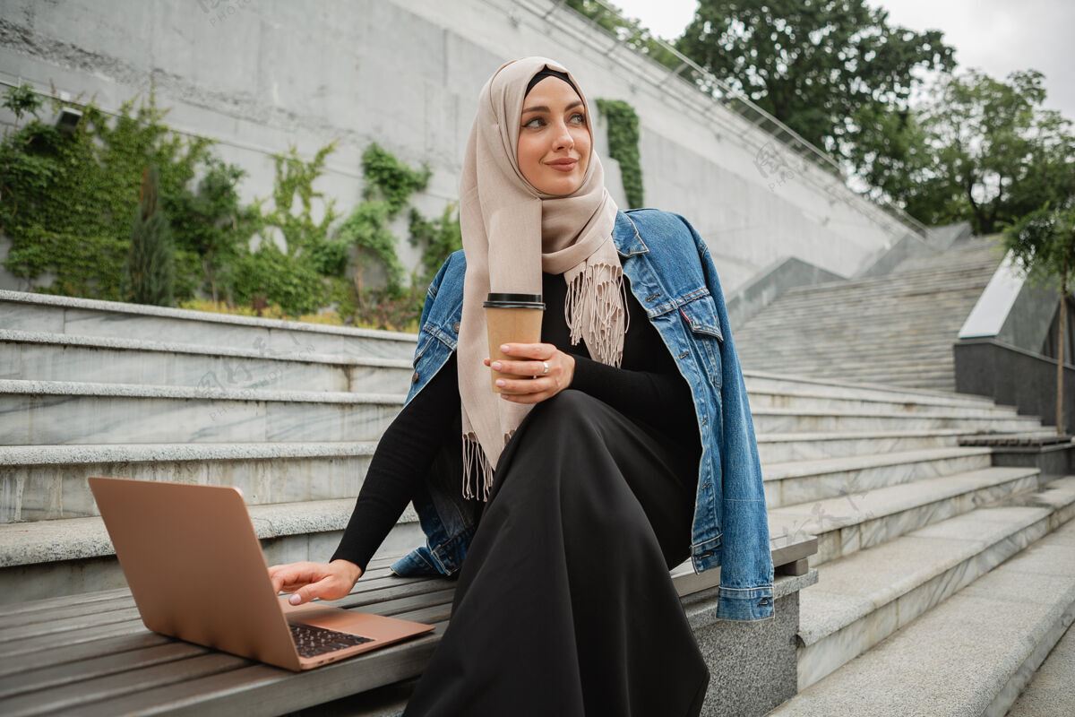 头巾现代时尚的穆斯林妇女戴着头巾 穿着牛仔夹克和黑色长袍坐在城市街道上用笔记本电脑工作迪拜服装女性