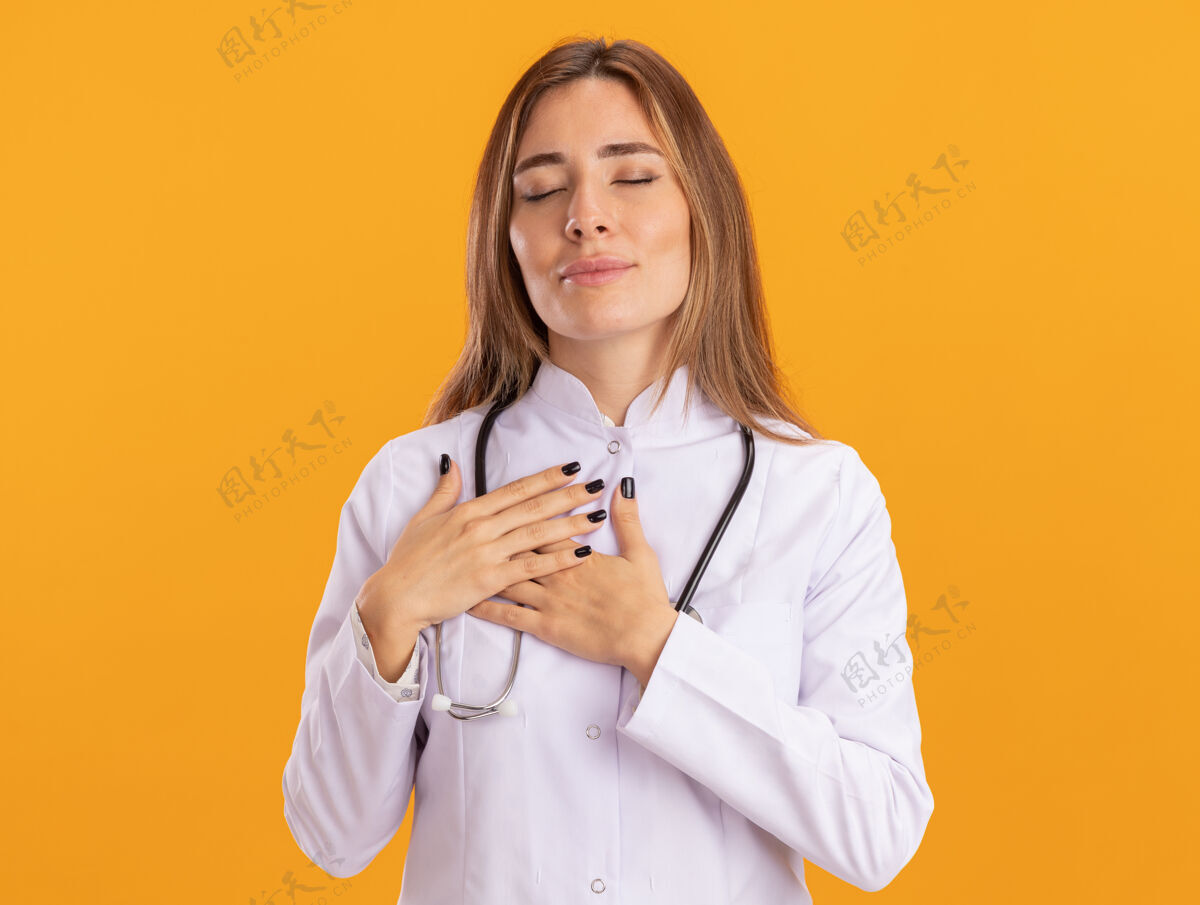 脸高兴地闭上眼睛的年轻女医生穿着医用长袍 用听诊器把手放在隔离在黄色墙上的心脏上长袍穿着人