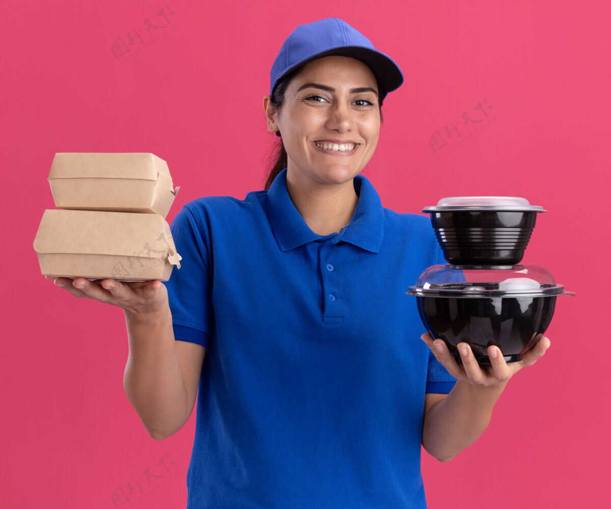 女孩带着微笑的年轻送货女孩穿着制服 戴着帽子 把食物容器隔离在粉红色的墙上公民站着食物