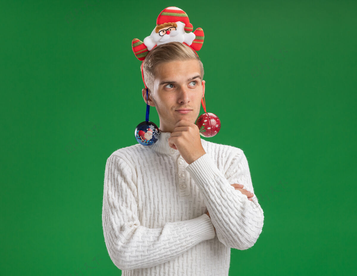 思想体贴的年轻帅哥戴着圣诞老人的头带抚摸着下巴看着一边 耳朵上挂着圣诞饰品 隔离在绿色的墙上 留着复制空间装饰家伙圣诞老人