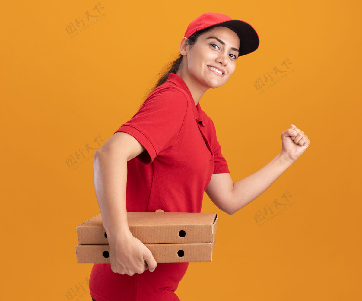 年轻人微笑着的年轻送货女孩穿着制服 戴着帽子 拿着披萨盒 展示着奔跑的姿态 隔离在橙色的墙上姿势感觉表演