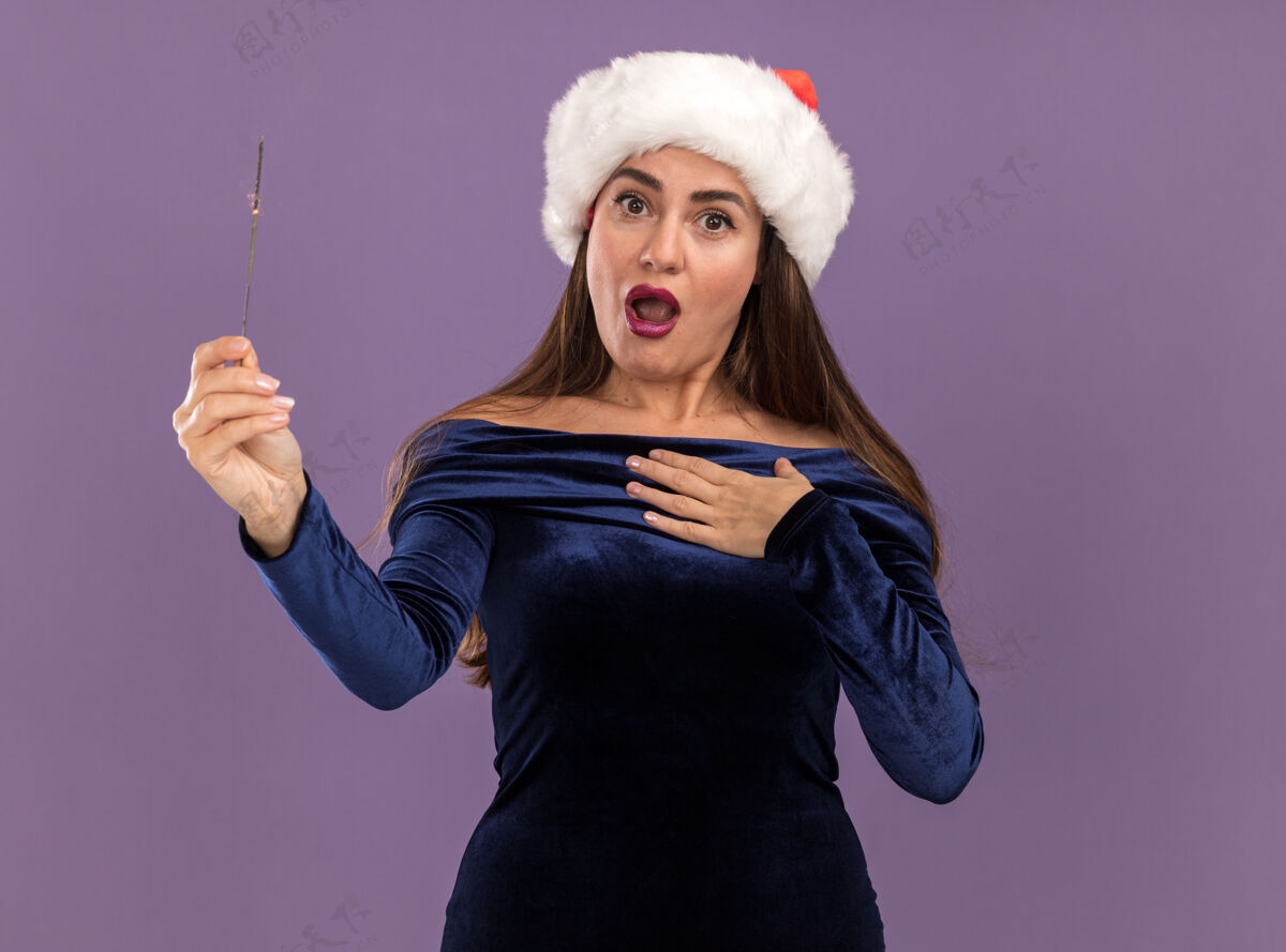 闪闪发光年轻漂亮的女孩穿着蓝色的裙子 戴着圣诞帽 手里拿着闪闪发光的灯 把自己孤立在紫色的墙上她自己穿害怕