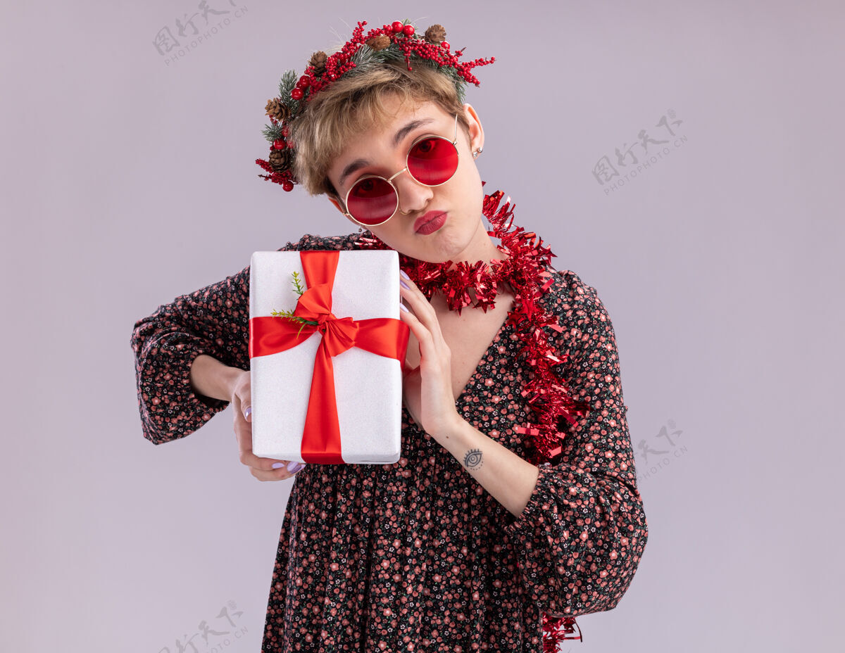 花环自信的年轻漂亮女孩戴着圣诞花环 脖子上戴着金箔花环 戴着眼镜 手里拿着礼物包 噘着嘴唇 隔离在白色墙壁上 留着复印空间女孩穿钱包