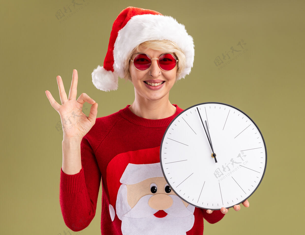橄榄微笑着的金发女郎 戴着圣诞帽 穿着圣诞老人的圣诞毛衣 戴着眼镜 拿着时钟 看起来很好 橄榄绿的墙上贴着一个孤立的牌子金发微笑女人