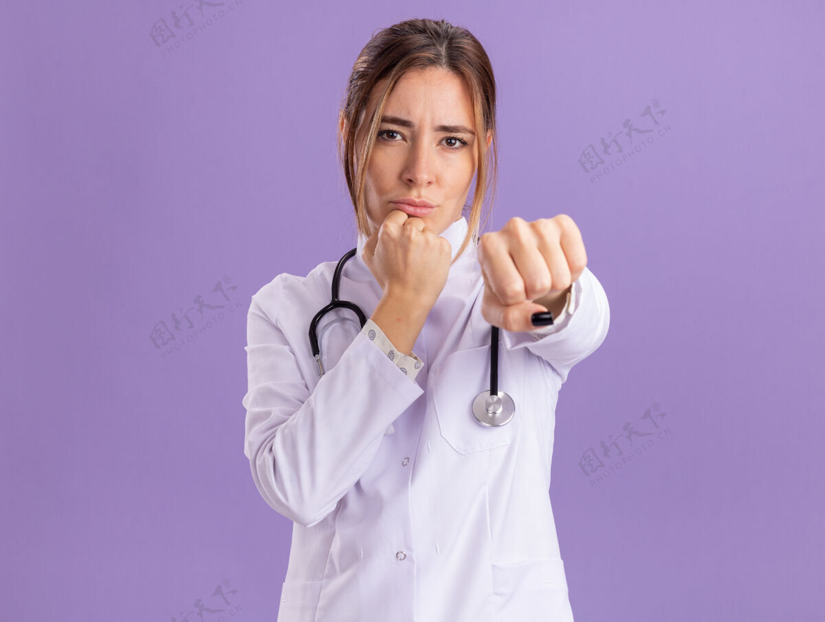 表情自信的年轻女医生穿着医用长袍 手持听诊器 对着隔离在紫色墙上的摄像机伸出拳头脸人人