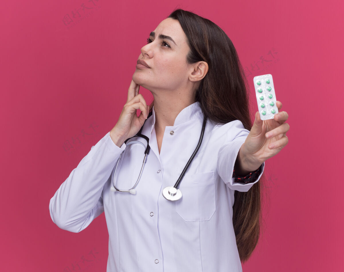 医学体贴的年轻女医生穿着带听诊器的医用长袍 手放在下巴上 手里拿着一包药 看着旁边包粉红色下巴