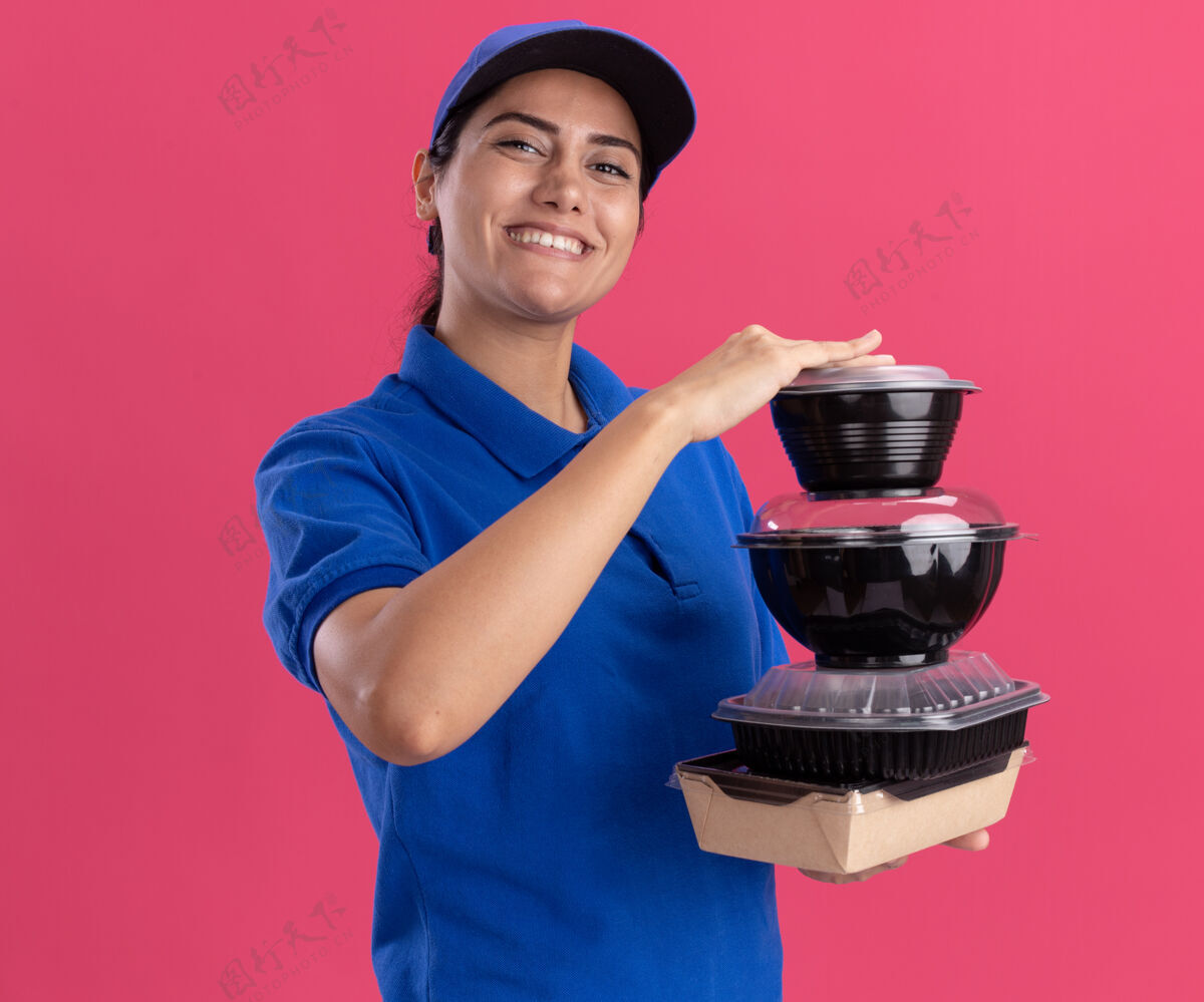 人带着微笑的年轻送货女孩穿着制服 戴着帽子 把食物容器隔离在粉红色的墙上人表情年轻人