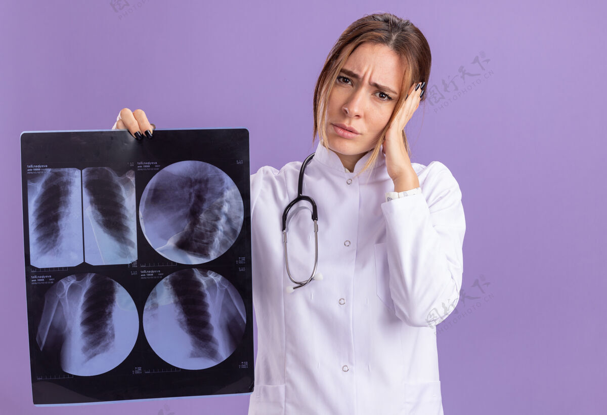 女性疲惫不堪的年轻女医生穿着医用长袍 手持听诊器 手持x光片 手放在隔离在紫色墙上的头上感觉人抱着