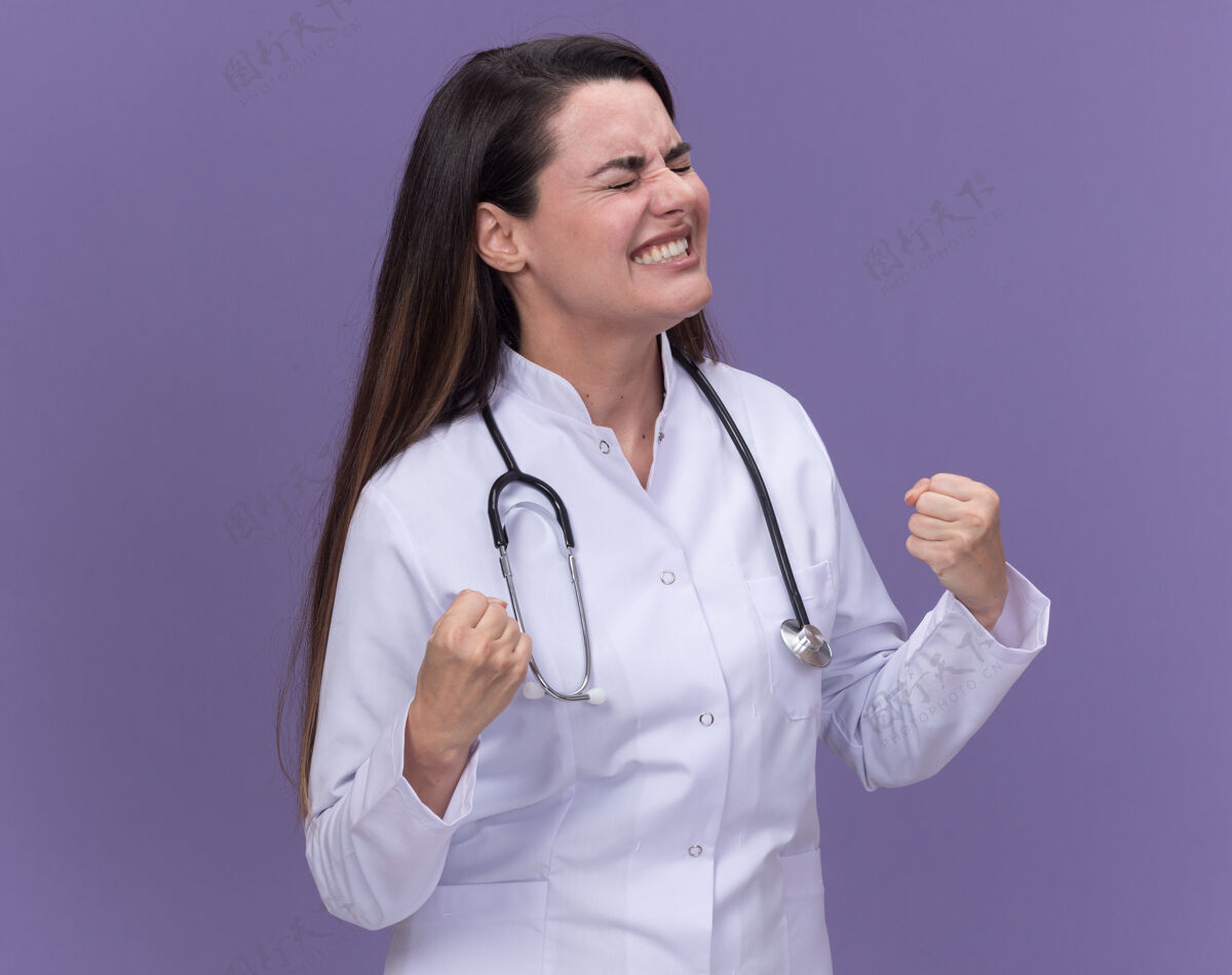保持兴奋的年轻女医生穿着医用长袍 手持听诊器 闭着眼睛站着 握紧拳头医生眼睛紫色