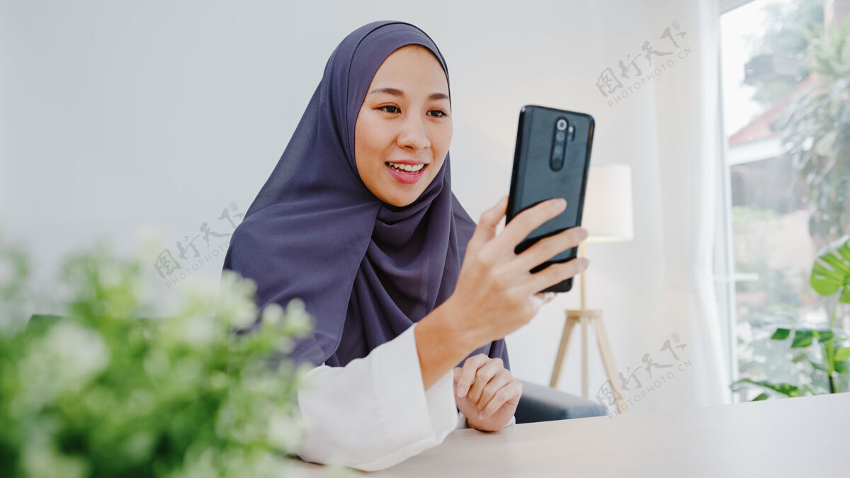 工作年轻的穆斯林女商人使用智能手机通过视频聊天与朋友交谈头脑风暴在线会议 同时在客厅远程工作网络团队青少年