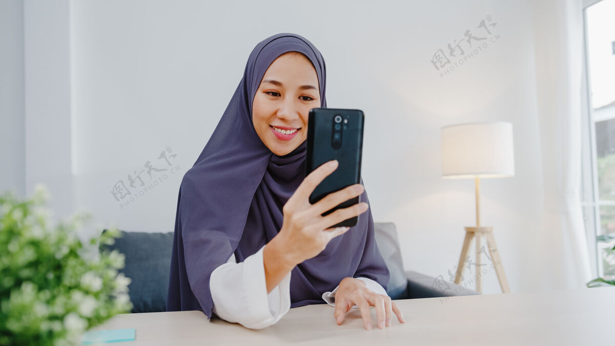 工作年轻的穆斯林女商人使用智能手机通过视频聊天与朋友交谈头脑风暴在线会议 同时在客厅远程工作办公室头巾虚拟