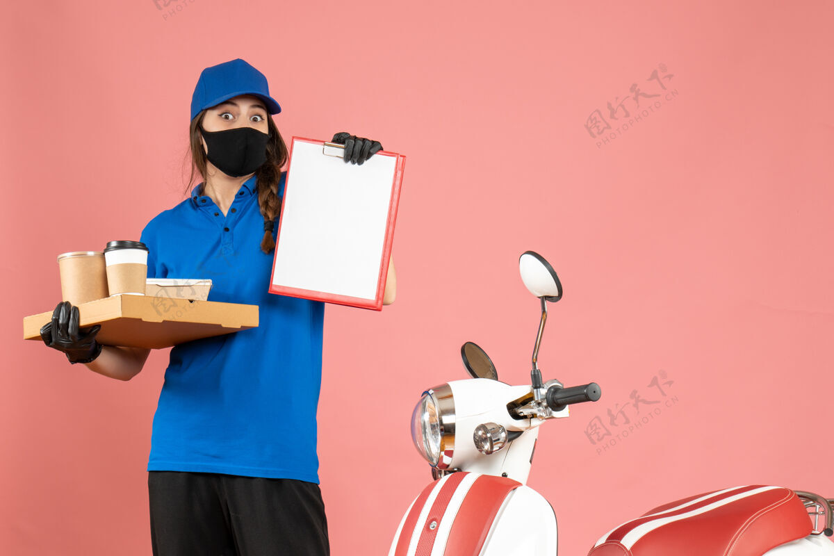 蛋糕俯视图：戴着医用口罩手套的信使女孩站在摩托车旁 手里拿着文件和咖啡小蛋糕 背景是柔和的桃色小站专业