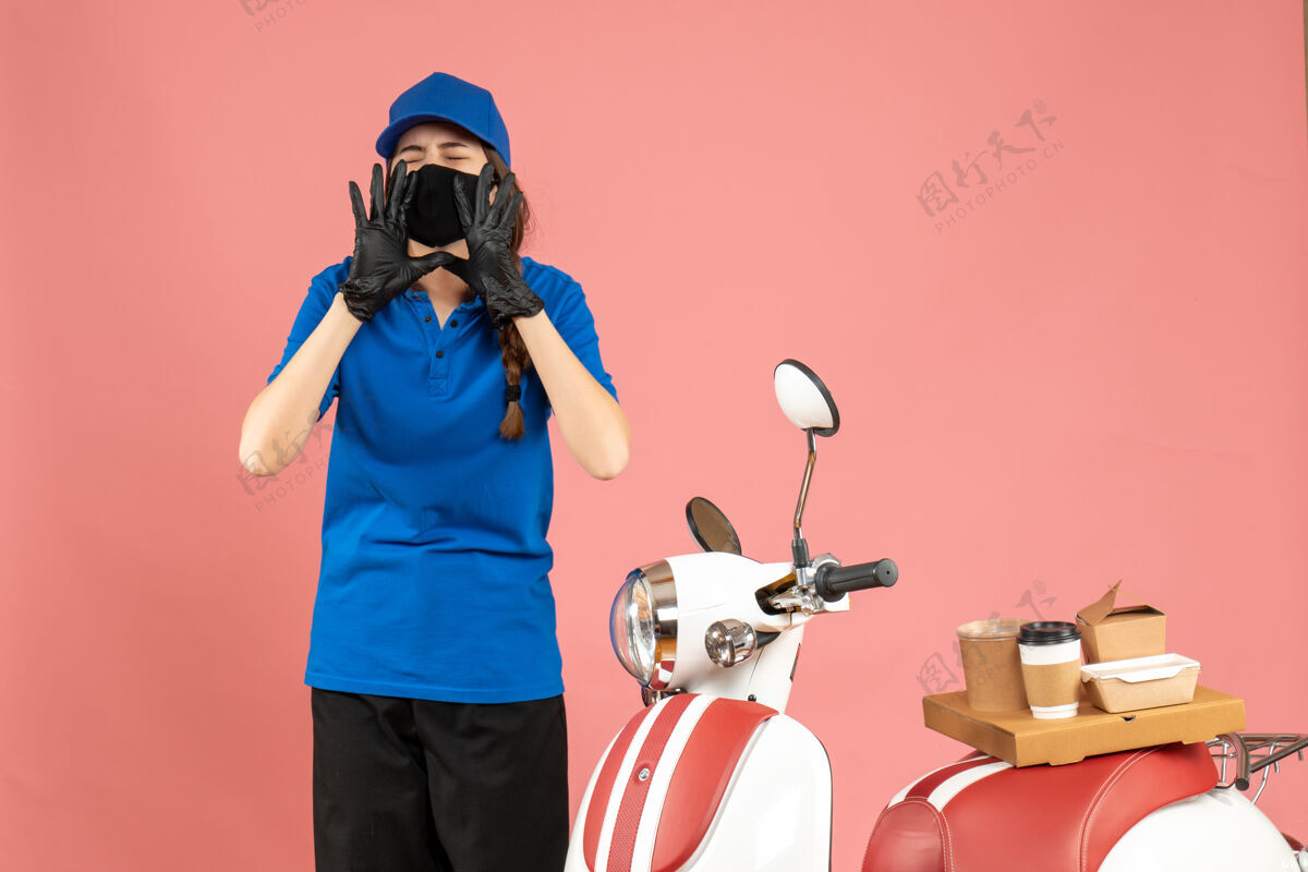 医药俯视图：戴着医用口罩手套的快递员女孩站在摩托车旁边 上面放着咖啡蛋糕 在柔和的桃色背景下呼唤着某人桃有人手套