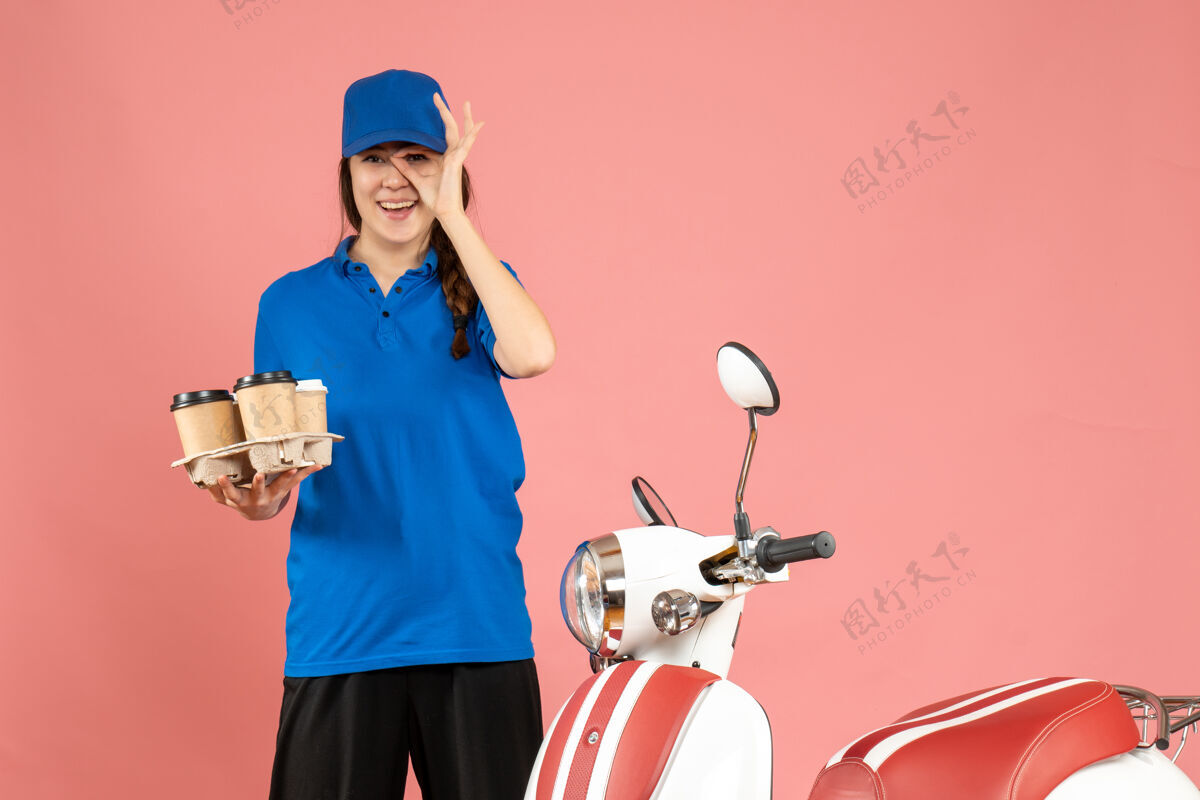 蛋糕正面图幸福微笑的信使女孩站在摩托车旁边拿着咖啡和小蛋糕在粉彩桃色背景上前面高尔夫站